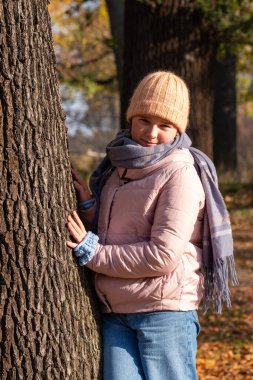 10 yaşındaki gülümseyen kız, sonbahar havasının tadını çıkarıyor, ağaçların yanında sarı yapraklarla duruyor. Açık havada aktif yürüyüşler.