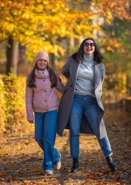 Anne ve kızı neşeyle gülüyor, el ele tutuşuyor, sonbahar parkında yürüyor. Aile mutluluğu, sonbahar huzuru. Kavram: Sevdiklerimizle geçirdiğimiz zamanın uyumu ve değeri.