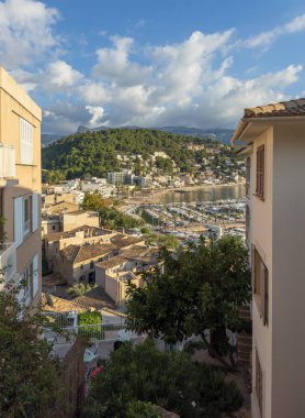 View across rooftops to the harbor of tourist town of Port de Soller: calm bay with yachts, surrounded by mountains, under bright Mediterranean sky clipart