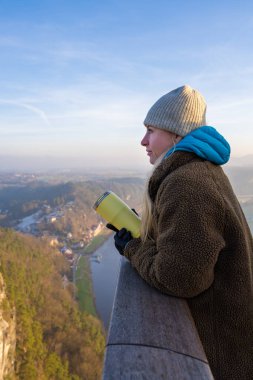 Young tourist in warm clothes with thermos in her hand stands on Bastai Bridge in Saxon Switzerland. Girl looks in distance at majestic cliffs, Elbe River.Popular tourist destination. Germany. clipart