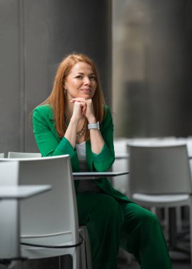 Yeşil takım elbiseli, kızıl saçlı güzel bir kadın açık hava kafede mola veriyor, açık hava masasında oturuyor. Her yerde kentsel bir atmosfer var. Profesyonellik, tarz ve rahat çalışma saatlerinin birleşimi. Boşluğu kopyala.