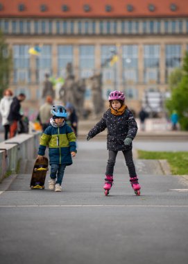 Girl is skating on roller skates, little boy is walking next to her with skateboard in his hands. Children having fun outdoors in cold season. clipart