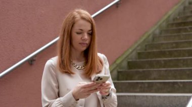 Genç kızıl saçlı kız akıllı telefondan numara çeviriyor, açık havada uzaktan konuşma yapıyor ve gülümsüyor. İletişim kavramı, şehir yaşam tarzı, modern teknoloji. 