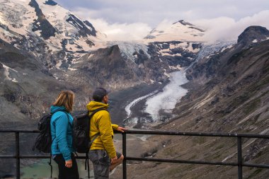 Middle aged man and woman stand on viewing platform overlooking Grossglockner Mountain and Pastertse Glacier in Hohe Tauern National Park, Austria. Concept: travel, outdoor activities. clipart