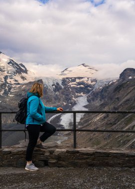 Middle aged woman stands on observation deck overlooking Grossglockner Mountain and Pasterze Glacier in Hohe Tauern National Park, Austria. Concept: travel, outdoor activities. clipart