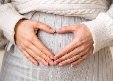Örgü örülü açık renkli elbiseli hamile kadının elleri kalp karnıyla bağlanmış. Yüzü olmadan. Bebek beklerken sağlıklı ve mutlu bir hamilelik..