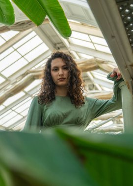 Yeşil yaprakların arasından kıvırcık saçlı kızın düşük açılı görüntüsü. Beyaz kız kameraya doğru bakıyor. Doğal güzellik kavramı ve doğa ile uyum.