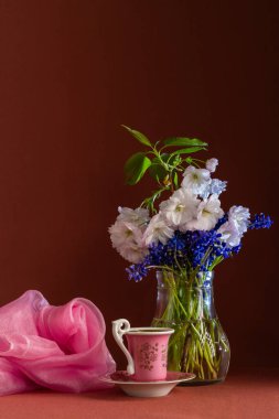 Pembe bardaklı, beyaz kiraz çiçeklerinden oluşan narin bir buket ve şeffaf vazoda mavi muscari çiçekleriyle dolu bir hayat, yanında koyu arkaplanda havadar bir şal yatar. Klasik stil