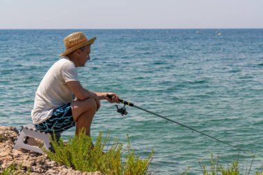 Şapkalı bir balıkçı elinde oltayla deniz kıyısında oturur. Yaz güneşli bir gün. Denizde sakin bir tatil, hobi.