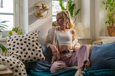 Sarışın, mutlu, genç bir kadın evdeki rahat koltukta oturup pencereden dışarı bakıyor. Rahat kadın portresi. Ev kıyafetleri içinde. Huzurlu bir anın tadını çıkarıyor. Ev konforu, kişisel bakım, akıl sağlığı kavramı.