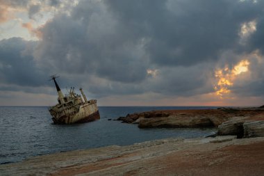 Kıbrıs 'ın kayalık kıyı şeridi yakınlarında terk edilmiş bir gemi enkazı manzarası. Sıcak altın ışık denizi yansıtıyor dramatik ve huzurlu deniz manzarası yaratıyor. Gizem ve seyahat kavramları için ideal.