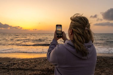 Orta yaşlı bir kadın kumsalda dururken akıllı telefonuyla günbatımını yakalıyor. Arkadan bak. Yumuşak altın ışık. Aşk kavramı, huzur, duygusal anlar ve anlamlı yalnızlık..