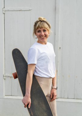 Beyaz ve bej elbiseli şık bir kadın, beyaz ahşap çitlerin yanında kaykay tutuyor. Enerji, özgürlük ve çağdaş şehir yaşam tarzını temsil eder..