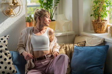 Gülümseyen genç sarışın kadın yumuşak kanepede dinleniyor ve dikkatlice pencereden dışarı bakıyor. Ev atmosferi, kişisel bakım ve zihinsel refah kavramı.