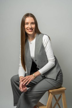 Modern stüdyoda zarif bir şekilde ahşap merdivende oturan, uzun dalgalı saçlı şık bir genç kadın tasviri. Basit ayar zarafeti ve özgüveni vurgular.