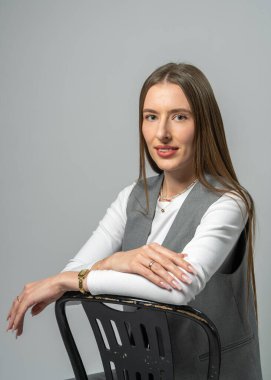 Koyu renk sandalyede oturan uzun saçlı hoş bir genç kadının stüdyo portresi. Tarafsız arka plan ve yumuşak ışıklandırma onun doğal cazibesini ve zarafetini vurguluyor..