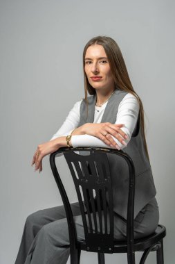 Uzun saçlı genç bir mankenin portresi. Temiz ve tarafsız arka planda koyu renkli bir sandalyeye oturmuş. Yumuşak stüdyo ışığı sıcaklık ve doğal cazibe ekler..