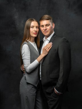 Klasik takım elbiseli zarif bir genç adam şık takım elbiseli çekici bir genç bayanın yanında poz veriyor. Stüdyo çekimleri koyu arkaplanlı. Çağdaş iş stilini ve modayı temsil eder..