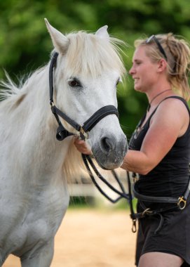 Beyaz atı nazikçe dizginleyen genç bir kadın, onu huzurlu kırsal alanda gezintiye hazırlıyor. İnsanlar ve hayvanlar arasındaki bağlantı kavramı, özen, güven.