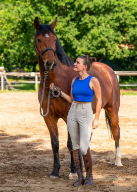 Kadın jokeyin yanında duran zarif safkan yarış atı. Kadın nazikçe atı tutuyor, ikisi de güneşli bir günde dışarıda poz veriyor. Binicilik sporu, at yarışı, hayvan arkadaşlığı kavramı..