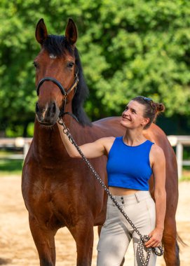 Güzel safkan yarış atı at çiftliğinde kadın jokeyin yanında duruyor. Açık havada güneşli bir gün. Jokey üniformalı kadın dizginleri elinde tutuyor, erkek ve hayvan arasındaki bağı gösteriyor.