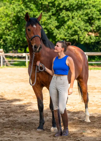 Kadın jokeyin yanında duran zarif safkan yarış atı. Kadın nazikçe atı tutuyor, ikisi de güneşli bir günde dışarıda poz veriyor. Binicilik sporu, at yarışı, hayvan arkadaşlığı kavramı..