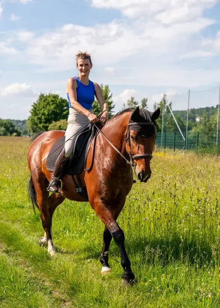Binicilik kıyafetleri giymiş kadın binici yeşil tarlalarda toprak yolda güzel bir yarış atı sürüyor. Yaz günü. Sahne, etkin bir yaşam tarzı, güç ve doğa ile uyumu simgeler..