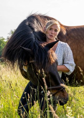 Açık arazide kahverengi atın yanında duran genç bir kadın. Huzurlu kırsal yerleşim, doğal ışık, hayvan dostluğu ve özgürlük kavramı.