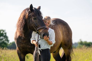 Genç bir kadın, güneşli çayırlarda dikilirken kahverengi atını sevgiyle okşar. Kırsal kesimde binici ve hayvan arasındaki güçlü bağı ve güveni yakalayan samimi an..