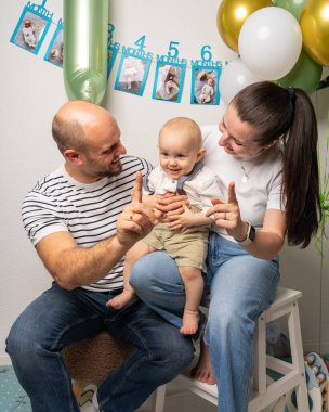 Anne ve baba bir yaşındaki oğullarını sevgiyle tutarken parmaklarıyla bir numarayı gösteriyorlar. Erken öğrenme, bağlantı ve çocukluk büyümesini vurgulayan tatlı ebeveynlik anı.