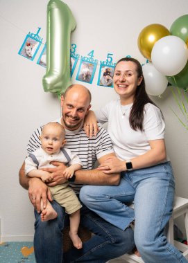 Mutlu ebeveynler oğullarını kucağına alıp parlak balonların yanında birlikte gülüyorlar. Yürek ısıtan aile sahnesi ilk doğum gününü aşk, neşe ve beraberlikle kutluyor..