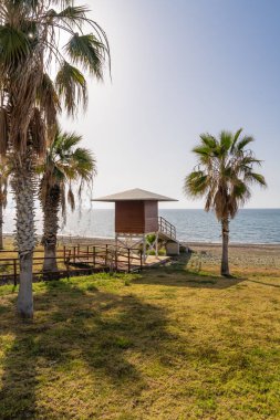 Güney Kıbrıs sahilindeki sakin Akdeniz manzaralı ahşap cankurtaran kulesinin palmiye ağaçları arasından manzara. Gün batımından önce. Seyahat, güvenlik, yaz veya tatil temaları için elverişli.