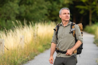 Orta yaşlı erkek yürüyüşçü doğa tarafından çevrili toprak yolda sırt çantasıyla tek başına yürüyor. Açık hava macerası, tek başına seyahat, kırsal yaşam tarzı, aktif tatil kavramı.