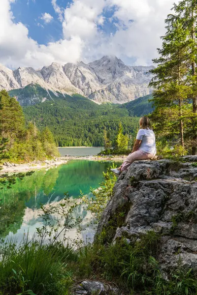 Eibsee Gölü ve Almanya 'daki Bavyera Alpleri' ne bakan kayada oturan kadın. Huzurlu alp manzarası, yürüyüş, doğa keşfi, seyahat ilhamı için ideal..