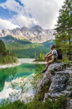 Taşın üzerinde oturan yürüyüşçü Eibsee Gölü 'nün berrak sularına ve Zugspitze yakınlarındaki dağlık tepelere bakıyor. Alman Alplerinde serin seyahat ve macera konsepti.