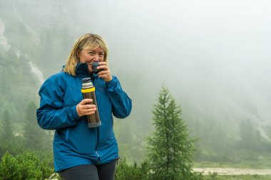 Sisli Dolomitler içinde yürürken termostan içen orta yaşlı bir kadın. Yağmurlu havada yağmurluk giyiyordu. Sakin ve kasvetli dağ atmosferi.