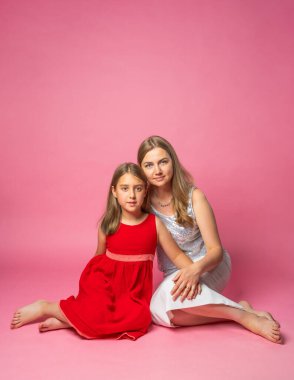 Kadın ve çocuk yerde otururken kucaklaşıyorlar, kırmızı ve beyaz giyinmiş, pembe arka planda. Ebeveyn-çocuk aşkı, şefkat, portre.