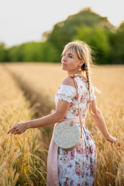 Bavyera tarzı örgülü, buğday tarlasında yürüyen ve yüzünü kameraya çeviren bir kız. Kırsal doğal ortamda kültürel portre.