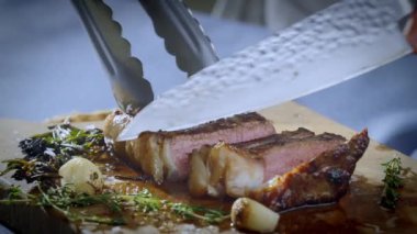 Aşçı kesme tahtasındaki büyük bir mutfak bıçağıyla bifteği kesiyor. Sulu biftek parçalara ayrıldı. Yavaş çekim videosu. Yüksek kalite 4k görüntü