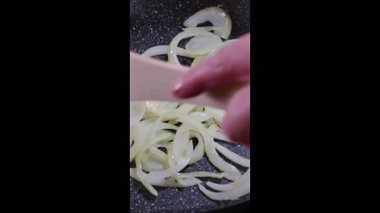 Dikey çekim. Yemek pişirmek. Kızarmış tavada doğranmış soğan. Aşçı ahşap bir spatulayla kızartma tavasındaki soğanı karıştırır. Seçici odaklanma. Yavaş çekim 50 fps.
