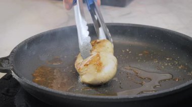 Aşçı sıcak tavada bir parça tavuk fileto pişiriyor, metal maşayla çeviriyor. Yüksek kalite 4k görüntü