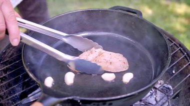 Bir şefin metal maşa kullanarak dışarıda kömürde pişen dökme demir bir tavada tavuk filetosu çevirdiği yakın plan. Yemek hazırlama. Yavaş çekim 100 fp. Yüksek kaliteli FullHD görüntüler