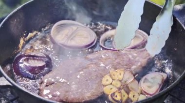 Yemek hazırlama. İyi pişmiş bir bifteğin kömürün üzerinde demir bir tavada pişirildiği ve bahçede ızgaranın üstünde ateş yakıldığı yakın plan. Aşçı bifteği ters çevirir. Yüksek kaliteli FullHD görüntüler