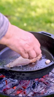 Dikey video. Açık havada ızgarada, dökme demir bir tavada zeytinyağında kızartılan sulu bir tavuk filetosunun yakın çekimi. Şef tavaya sarımsak koyar. Yüksek kaliteli FullHD görüntüler