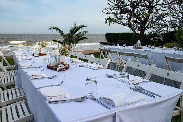 Table dinner set ready for an outdoor party near the beach. White tone decoration with white wooden chairs set.
