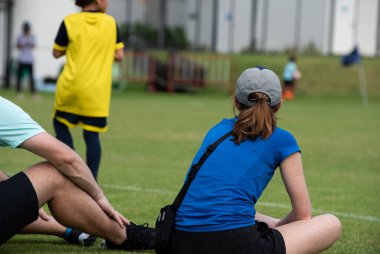 Anne, güneşli bir günde okul futbol turnuvasında oturup oğlunun futbol oynamasını izliyor. Spor, açık hava etkinliği, yaşam tarzı, mutlu aile ve futbol annesi ve futbolcu baba kavramı..