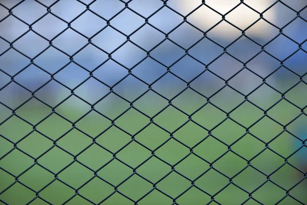 A close-up of a chain link fence in a sport field with a nice scene of a football field and sport hub as a background in the evening.