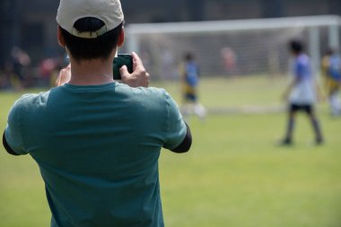 Futbolcu baba oğlunun açık havada ve güneşli bir günde okul turnuvasında futbol oynarken videosunu çekiyor. Spor, aktif yaşam tarzı, mutlu aile ve ev hanımı anne ve baba kavramı..