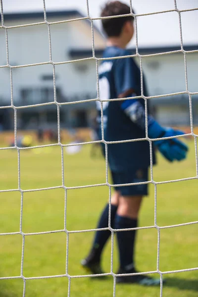 Yakın plan futbol kalesi, genç bir futbolcuyu takım arkadaşının maçını izlerken görmek. Güneşli bir yaz okulu turnuvasında gençler futbol maçı. Arka planda devam eden bir futbol maçı.