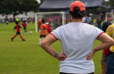 Anne, güneşli bir günde okul futbol turnuvasında oğlunun maçını seyrediyor. Spor, açık hava etkinliği, yaşam tarzı, mutlu aile ve futbol annesi ve futbolcu baba kavramı..
